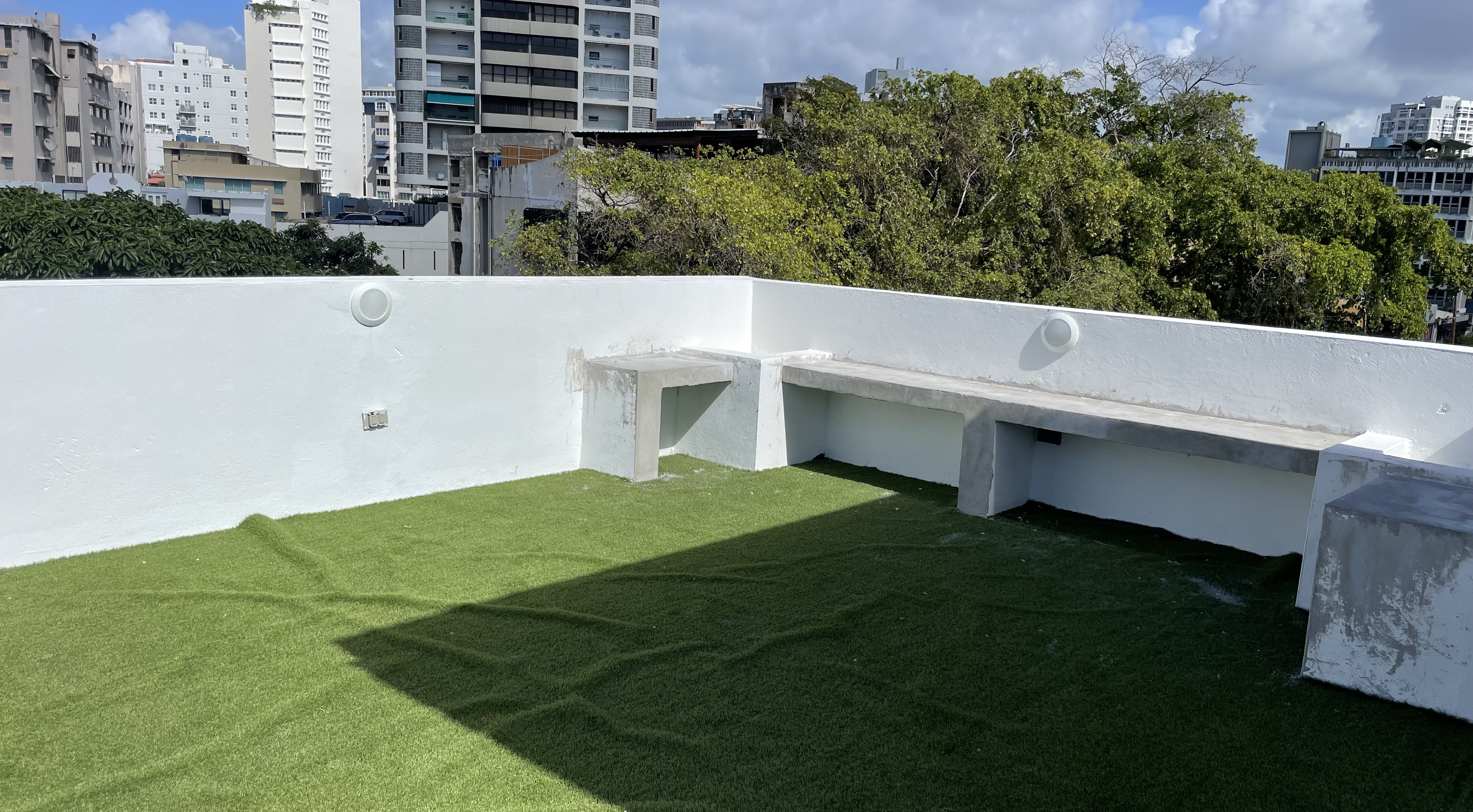 Terraza moderna en una azotea con césped artificial verde, bancos de concreto integrados y una pared blanca baja, con vistas a edificios urbanos y árboles frondosos de fondo