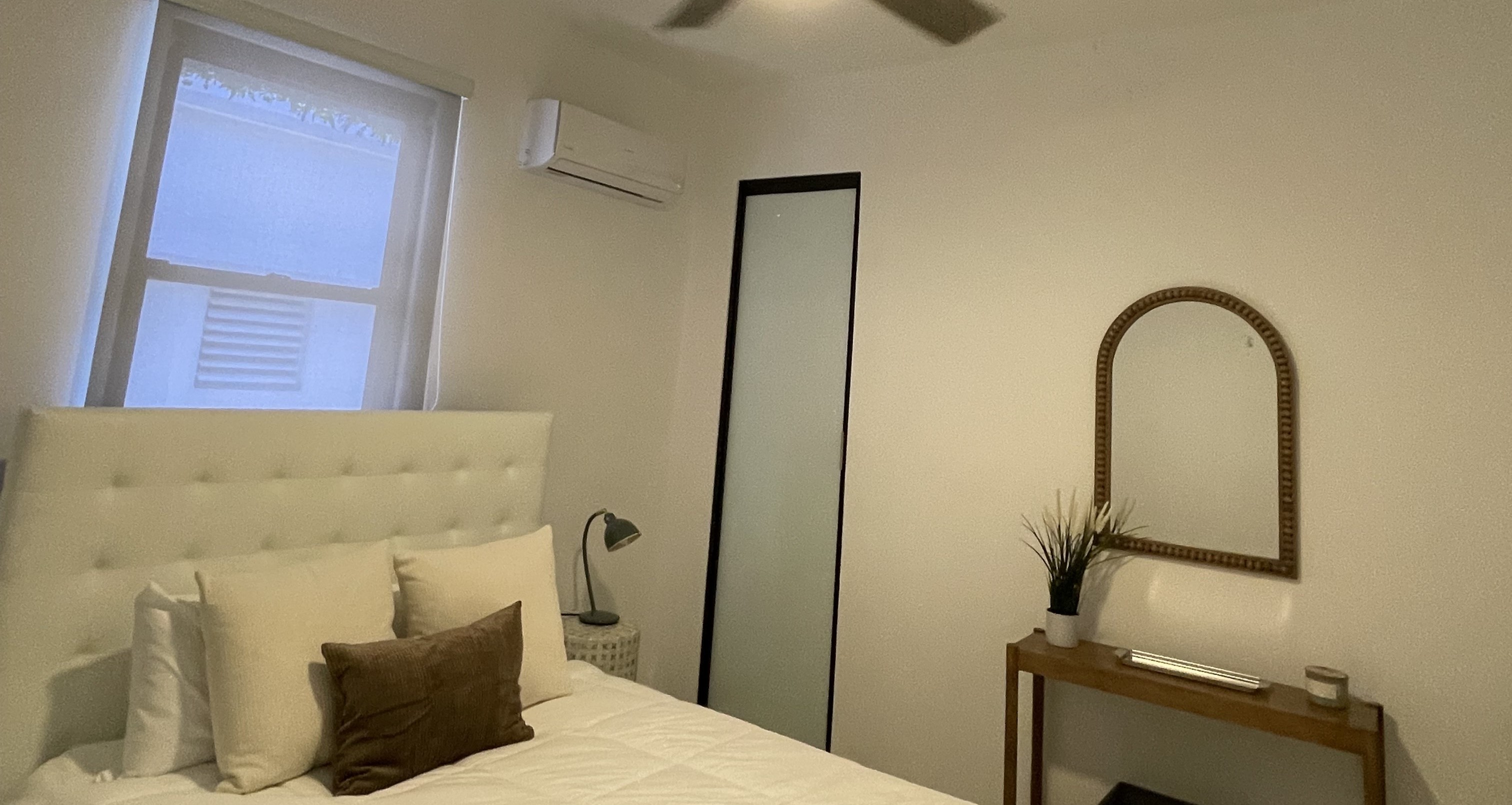 Dormitorio moderno con cama blanca de cabecero acolchado, aire acondicionado de pared, una consola de madera con espejo arqueado y una ventana con luz natural suave