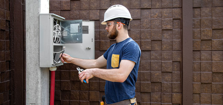 Electricista con casco de seguridad y gafas protectoras realizando mantenimiento en un panel eléctrico exterior montado en un poste