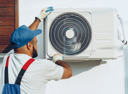 Técnico instalador con gorra azul y guantes ajustando una unidad externa de aire acondicionado split