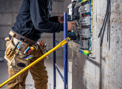 Electricista profesional utilizando un taladro inalámbrico para trabajar en un panel de interruptores automático dentro de una construcción.
